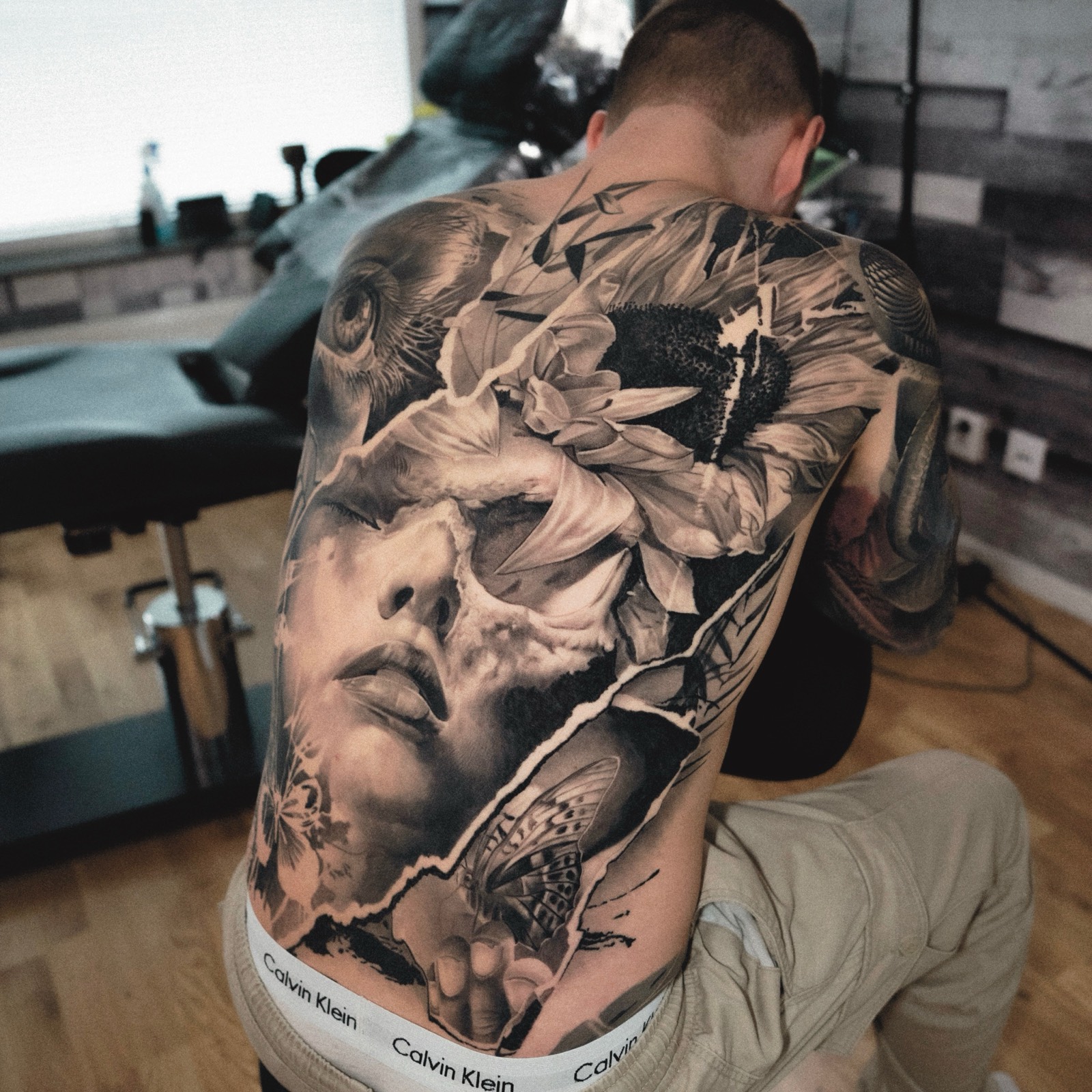 Black and grey back piece blending a portrait, skull, sunflower and butterfly
